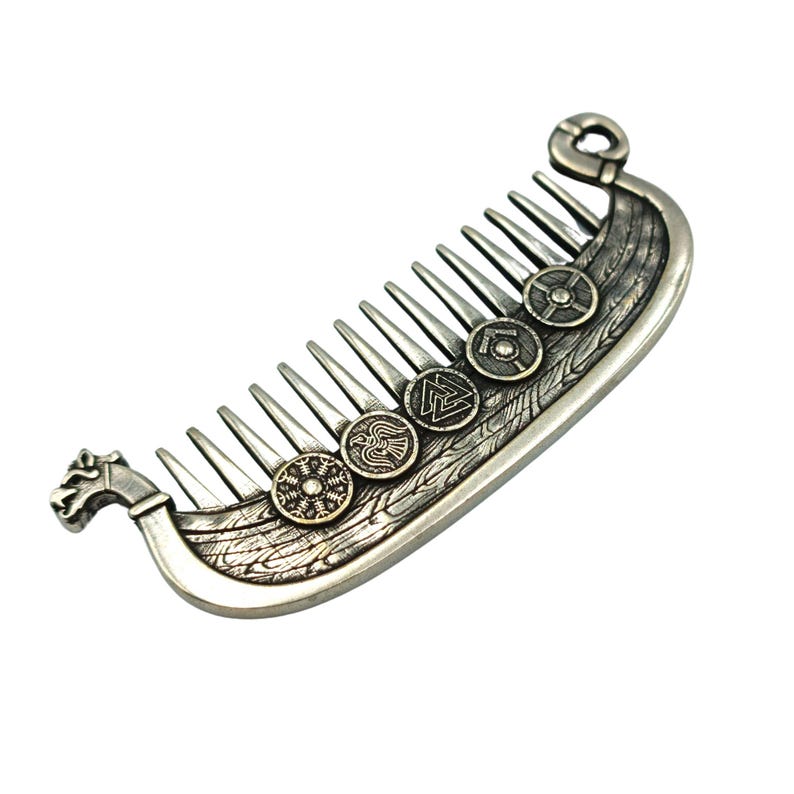 A silver-colored comb shaped like a Viking ship Drakkar, featuring intricate details and decorative round emblems. The comb's teeth are evenly spaced, and the ship design includes a dragon head at the front. The emblems have various symbols.