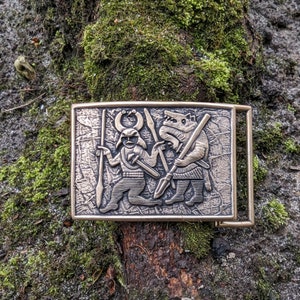 May include: A rectangular bronze-toned belt buckle featuring a raised relief design of a horned figure holding a spear and a sword, standing beside a wolf-like creature holding a spear. The metal surface has a textured, aged finish.