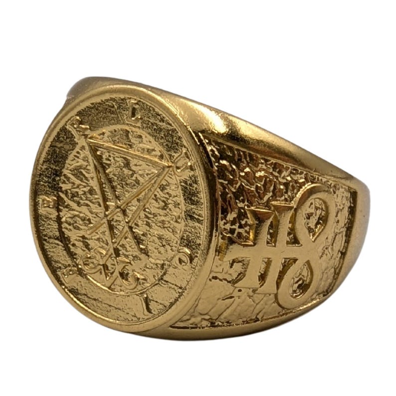 May include: A gold-toned ring featuring a circular face with an engraved sigil of Lucifer and the letters L, U, C, I, F, E, R arranged around the perimeter. The side of the band displays a raised, textured finish with an additional embossed symbol.