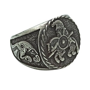 May include: A silver ring with a detailed design of a warrior on horseback. The warrior is wearing a helmet and holding a spear. The ring is set against a white background.
