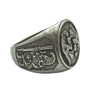 May include: A silver ring with an intricate design featuring a figure holding a rifle and a bird with outstretched wings.