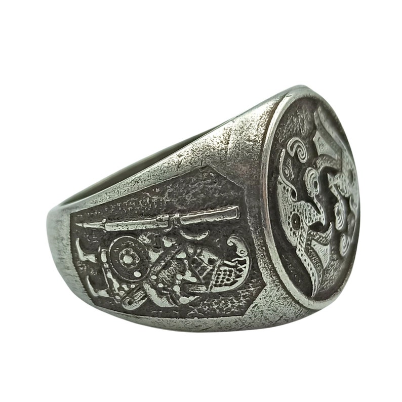 May include: A silver ring with an intricate design featuring a figure holding a rifle and a bird with outstretched wings.