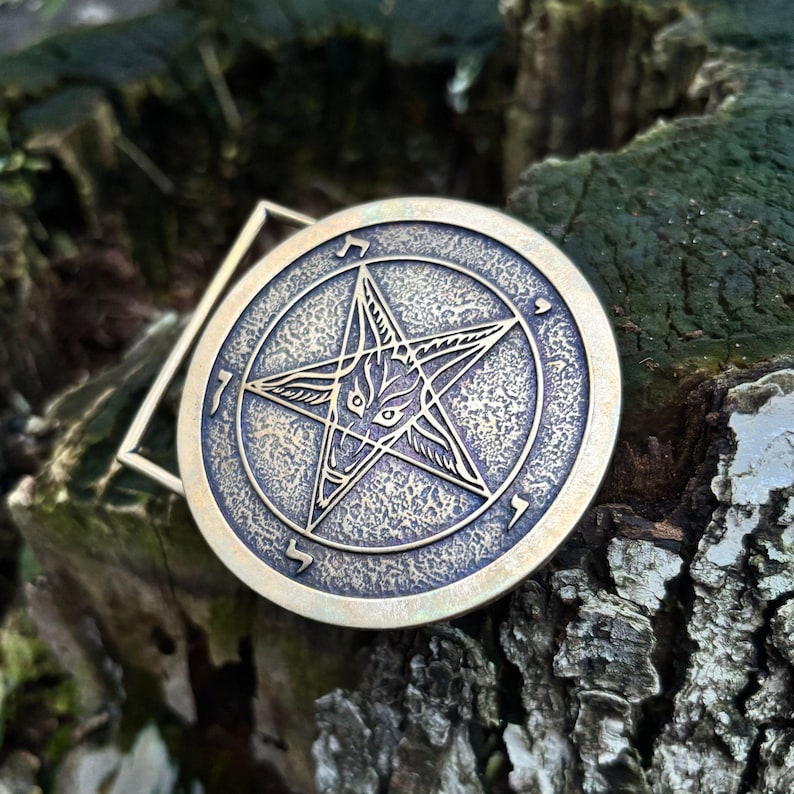 May include: A circular brass-toned metal belt buckle featuring an embossed pentagram design with a central goat head motif. The surface has a textured, antiqued finish with Hebrew characters engraved along the inner circular border of the emblem.