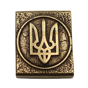 May include: A gold-colored metal square with a raised, circular design featuring the Ukrainian trident symbol. The trident is a stylized three-pronged spear, a national symbol of Ukraine.