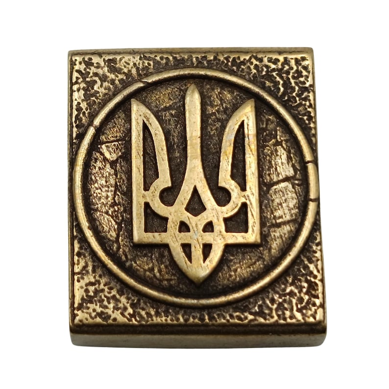 May include: A gold-colored metal square with a raised, circular design featuring the Ukrainian trident symbol. The trident is a stylized three-pronged spear, a national symbol of Ukraine.