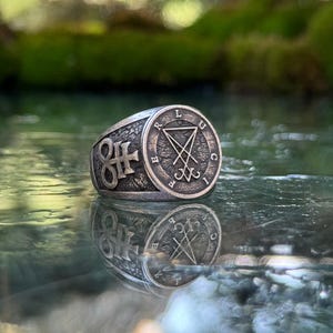 May include: A silver-toned metal ring with a textured, dark finish. The circular face features a central sigil of Lucifer symbol enclosed by the letters L, U, C, I, F, E, R. The side of the band displays a prominent Leviathan cross symbol.