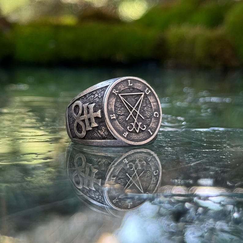 May include: A silver-toned metal ring with a textured, dark finish. The circular face features a central sigil of Lucifer symbol enclosed by the letters L, U, C, I, F, E, R. The side of the band displays a prominent Leviathan cross symbol.