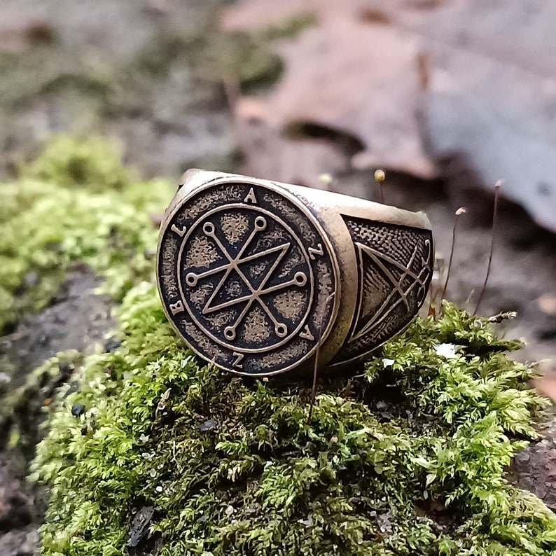 A bronze ring with a circular design featuring a Azazel sigil ring. The ring has a textured band with a smaller design on the side.