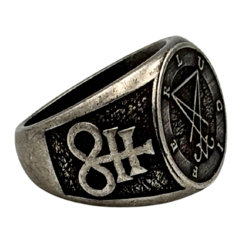 May include: A silver-toned metal ring with a dark, oxidized finish. The band features a prominent symbol resembling a stylized infinity loop with a cross. A circular face displays the sigil of Lucifer with the text LUCIFER inscribed around the edge.