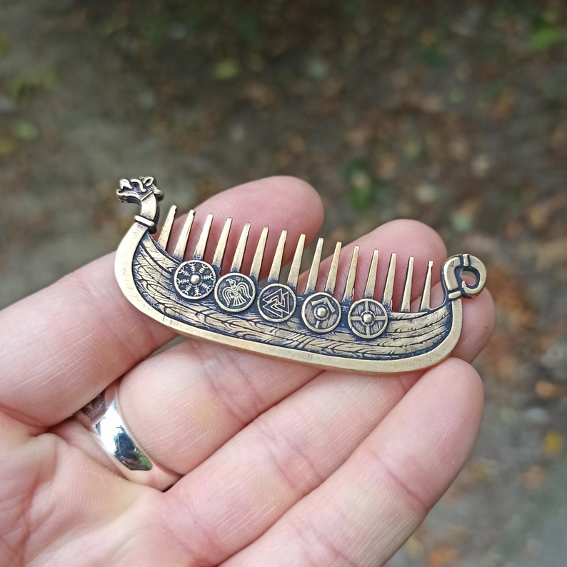 May include: A bronze metal beard Viking ship Drakkar comb with intricate detailing. The comb features a ship design with a dragon head at the front and a decorative loop at the back. The comb's teeth are vertical and the ship's body has circular shield designs.