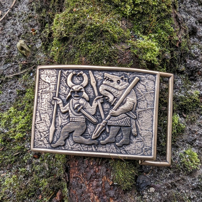 May include: A rectangular bronze-toned belt buckle featuring a raised relief design of a horned figure holding a spear and a wolf-like creature holding a spear. The metal surface has a textured, weathered finish with a rectangular frame and loop.