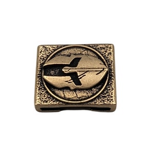 May include: A gold-colored metal square tile with a raised circular design. The circle features a stylized image of a spaceship in black.