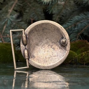 May include: A circular belt buckle made of aged brass with a textured, mottled surface. The buckle features a rectangular frame attached to the side, designed to hold a belt strap, and includes a small metal post on the back for secure fastening.