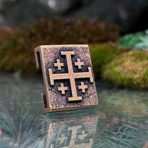 May include: A rectangular bronze-toned metal bead featuring a raised Jerusalem cross design on its front surface. The metal has a dark, weathered, and textured finish with visible cracks and aged detailing across the entire face of the piece.