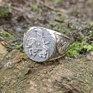 May include: A silver ring with a detailed design of a rider on a horse with a shield. The ring is set against a background of moss and bark.