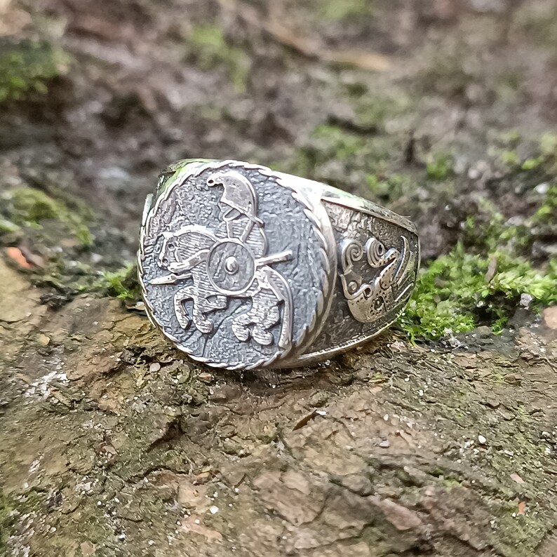 May include: A silver ring with a detailed design of a rider on a horse with a shield. The ring is set against a background of moss and bark.