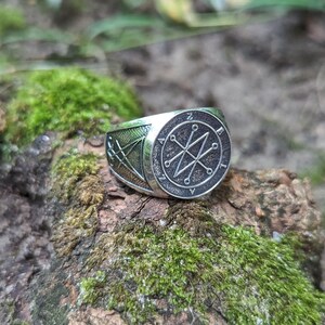 A silver ring with a Azazel sigil design featuring a star with six points and a circle around it. The ring is set on a mossy green surface.