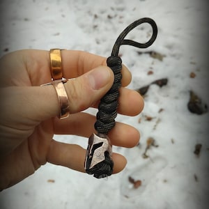 May include: A black paracord keychain with a copper Spartan helmet charm. The keychain is held in a hand wearing two gold rings.