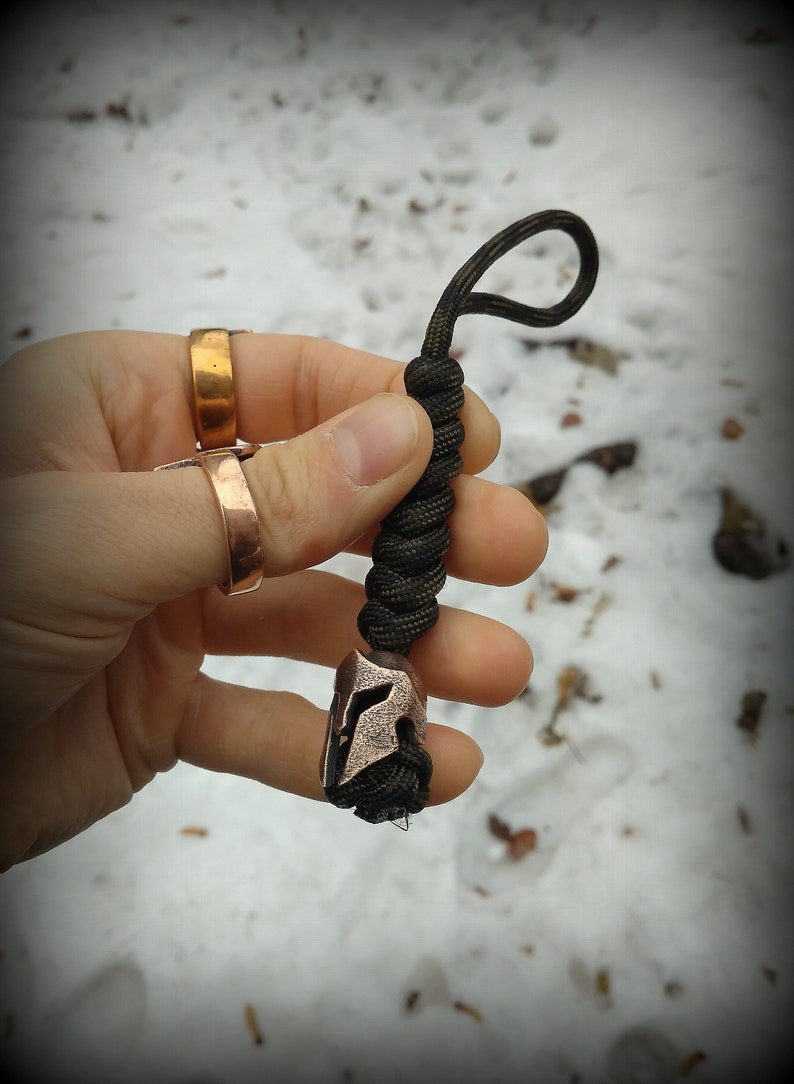 May include: A black paracord keychain with a copper Spartan helmet charm. The keychain is held in a hand wearing two gold rings.