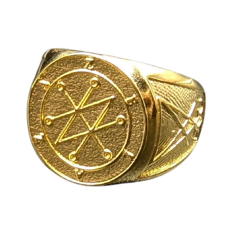 A gold ring with a circular design featuring a starburst pattern and a geometric symbol in the center - the Azazel sigil.