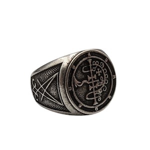 May include: A silver ring with a circular design featuring a sigil and symbols. The ring has a textured band with a triangular design.