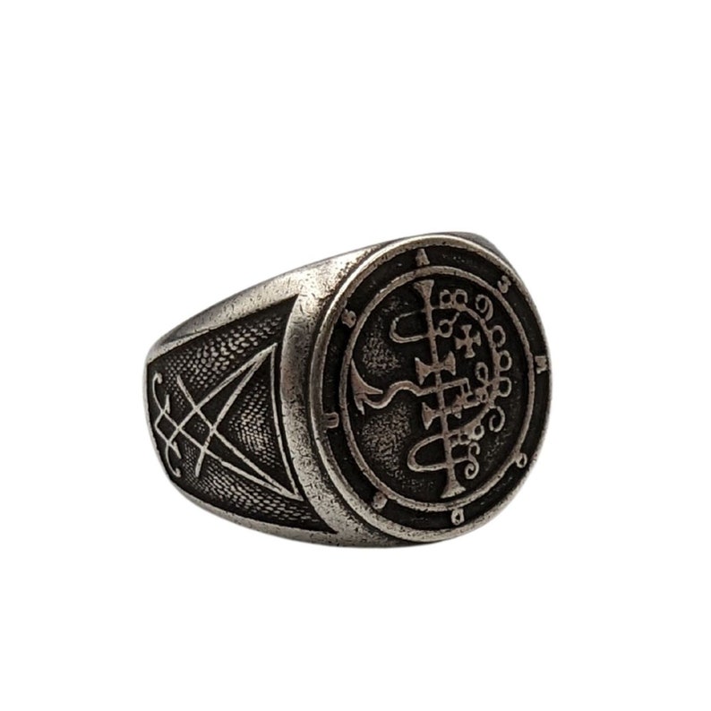May include: A silver ring with a circular design featuring a sigil and symbols. The ring has a textured band with a triangular design.