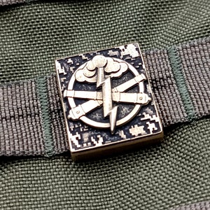 Op de afbeelding: Een gouden metalen patch met een zwart-wit camouflagepatroon. De patch heeft een bliksem, een wolk en drie gekruiste kanonnen.