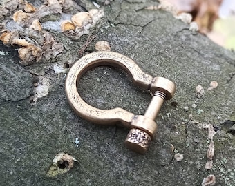 May include: A brass U-shaped clasp with a threaded screw. The clasp has a textured surface and is lying on a piece of bark.