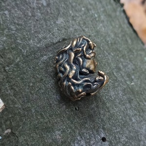 Puede incluir: Un colgante de bronce con una escultura detallada de la cara de una mujer con serpientes en el pelo.