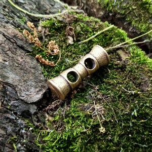 May include: A brass whistle with two holes, lying on a bed of green moss. The whistle is cylindrical in shape with a rounded top and bottom.