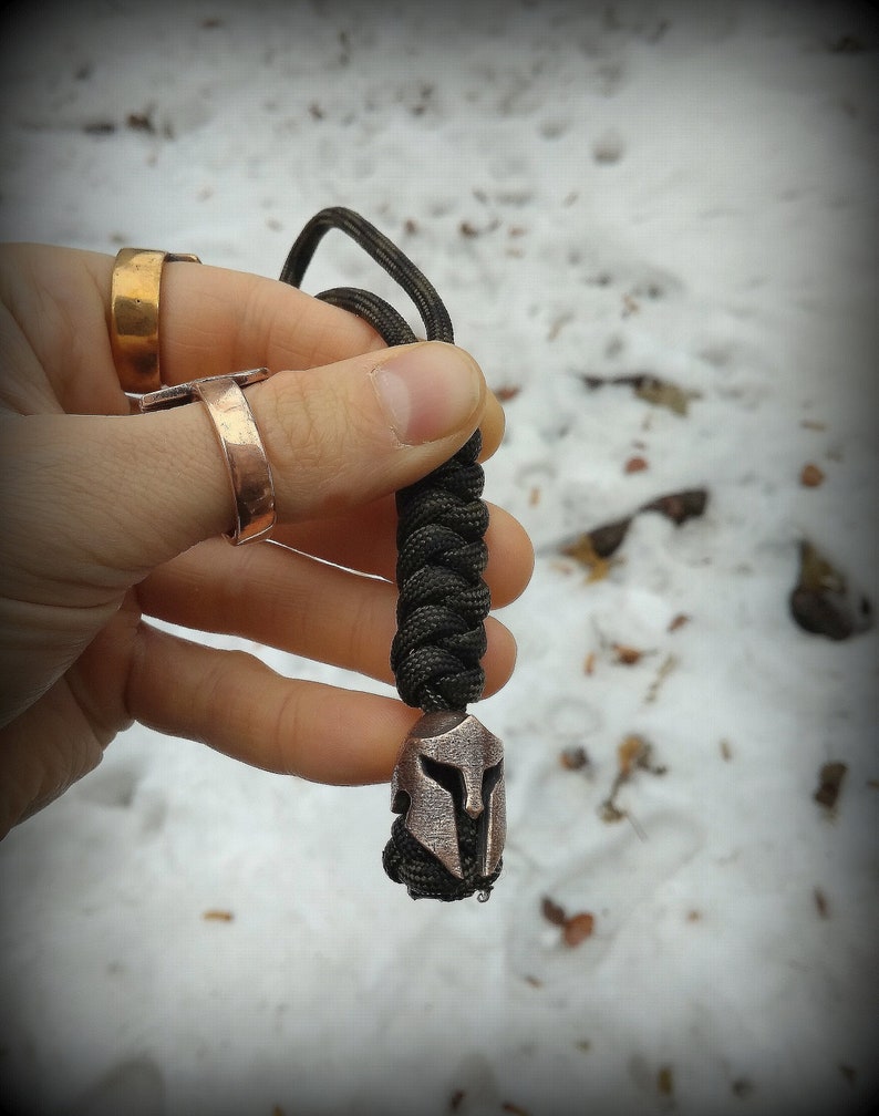 May include: A black paracord keychain with a silver Spartan helmet charm. The keychain is held in a hand wearing two rings.