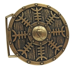 May include: A gold-toned belt buckle with a round design. The buckle features a circular shield with a wood grain texture and a central boss. The shield is decorated with a repeating pattern of stylised branches.