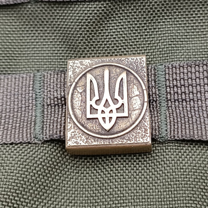 May include: A close-up of a metal badge with a gold finish. The badge features a stylized trident, the national symbol of Ukraine, in a circular design.