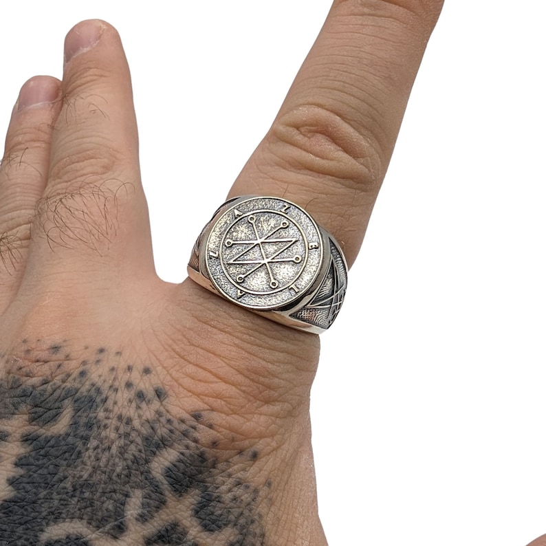 A silver ring with a circular design featuring a Azazel sigil. The ring has a textured band.