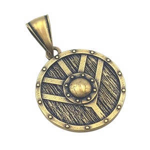 May include: A gold-toned metal pendant in the shape of a round shield with a raised centre and a textured surface. The shield has a decorative design of lines and a circle in the centre.