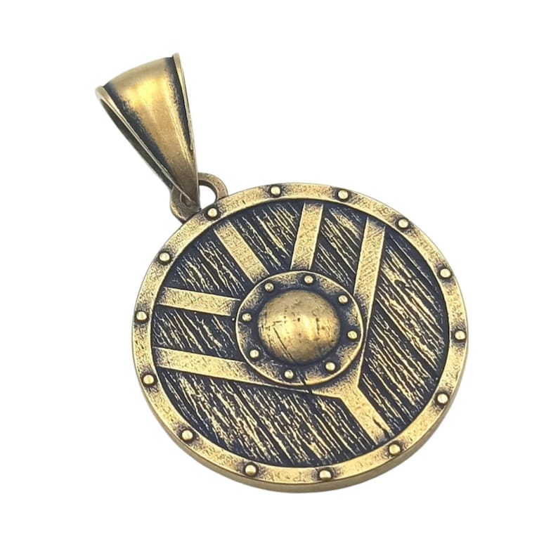 May include: A gold-toned metal pendant in the shape of a round shield with a raised centre and a textured surface. The shield has a decorative design of lines and a circle in the centre.