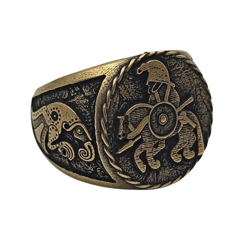 May include: A gold-toned ring with a detailed design of a warrior riding a chariot on the face of the ring. The ring is made of metal and has a textured finish.