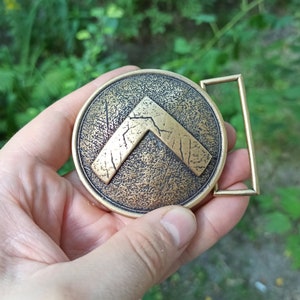 May include: A circular brass belt buckle featuring a raised, distressed lambda symbol in the centre. The surface of the buckle has a textured, weathered finish with dark patina accents, and it includes a rectangular loop on the right side.