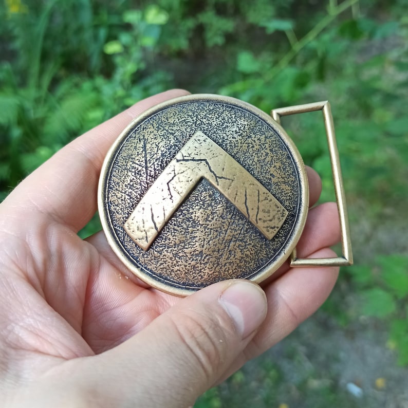 May include: A circular brass belt buckle featuring a raised, distressed lambda symbol in the centre. The surface of the buckle has a textured, weathered finish with dark patina accents, and it includes a rectangular loop on the right side.