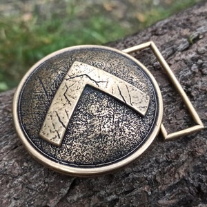 May include: A circular bronze-toned belt buckle featuring a raised, weathered Lambda symbol in the centre. The surface of the buckle has a textured, distressed finish with dark patina accents, and it includes a rectangular metal loop attachment.