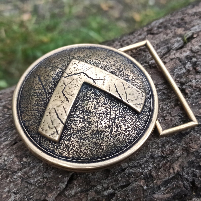 May include: A circular bronze-toned belt buckle featuring a raised, weathered Lambda symbol in the centre. The surface of the buckle has a textured, distressed finish with dark patina accents, and it includes a rectangular metal loop attachment.