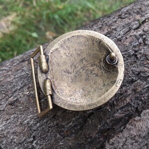 May include: A circular brass belt buckle with a weathered, antique finish. The reverse side features a rectangular loop attachment on the left side and a small, rounded metal stud on the right side, designed to secure the buckle to a belt strap.