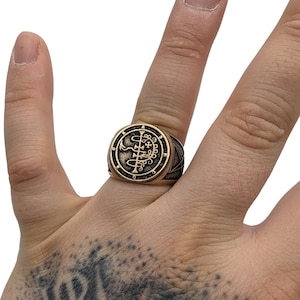 May include: A gold-toned ring with a black sigil design. The sigil is a complex symbol with a cross and other geometric shapes. The ring is worn on a person's finger.
