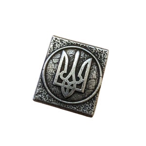 May include: A silver-colored metal pin with a textured surface. The pin features a raised, stylized image of the Ukrainian trident, a three-pronged spear.
