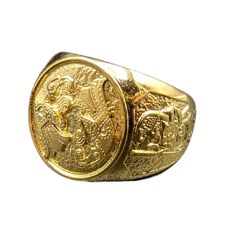 May include: A gold ring with an intricate design featuring a circular center piece with a detailed pattern and a textured band.