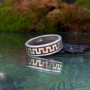 May include: A silver band ring featuring a continuous black engraved Greek key pattern around the exterior. The metal surface has a slightly weathered finish, and the ring is positioned horizontally, creating a clear reflection on the surface below.