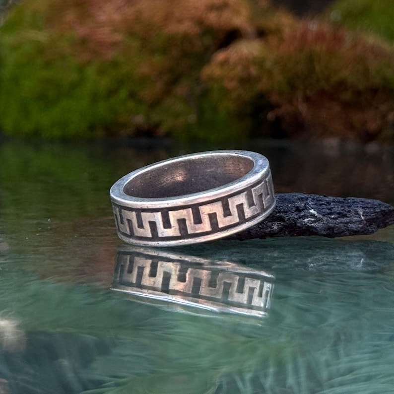 May include: A silver band ring featuring a continuous black engraved Greek key pattern around the exterior. The metal surface has a slightly weathered finish, and the ring is positioned horizontally, creating a clear reflection on the surface below.