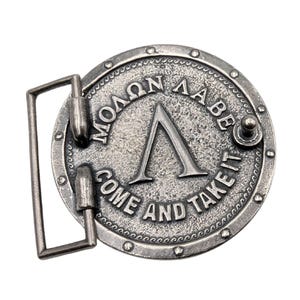 May include: A circular silver-toned metal belt buckle featuring a raised Greek letter Lambda in the centre. The text "&Mu;&Omicron;&Lambda;&Omega;&Nu; &Lambda;&Alpha;&Beta;&Epsilon;" is embossed above the letter, and the English text "COME AND TAKE IT" is embossed below, surrounded by a studded border.