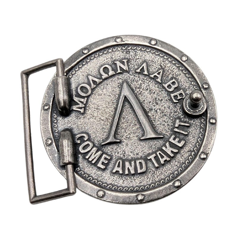 May include: A circular silver-toned metal belt buckle featuring a raised Greek letter Lambda in the centre. The text "&Mu;&Omicron;&Lambda;&Omega;&Nu; &Lambda;&Alpha;&Beta;&Epsilon;" is embossed above the letter, and the English text "COME AND TAKE IT" is embossed below, surrounded by a studded border.