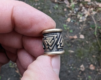 Op de afbeelding: Een gouden metalen kraal met een zwart en gouden ontwerp van in elkaar grijpende driehoeken. De kraal wordt in een hand gehouden.